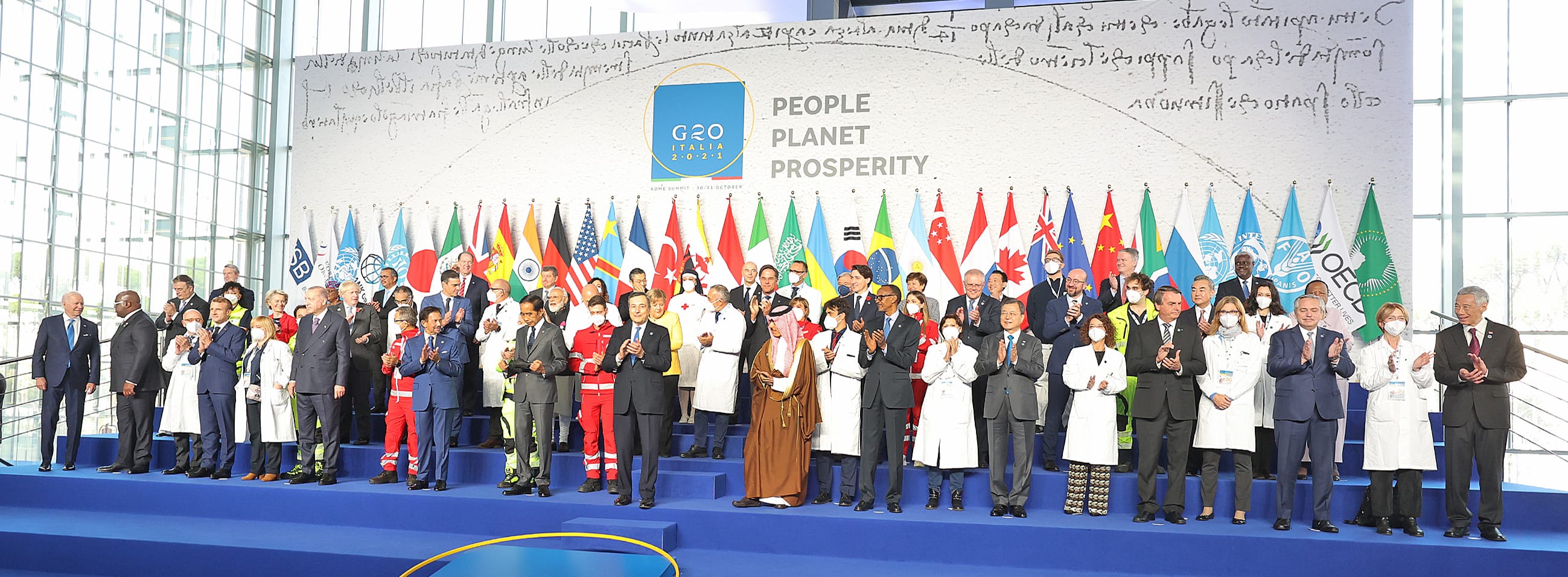G20 summit photo: Group of leaders standing below flags, clapping on blue stage.